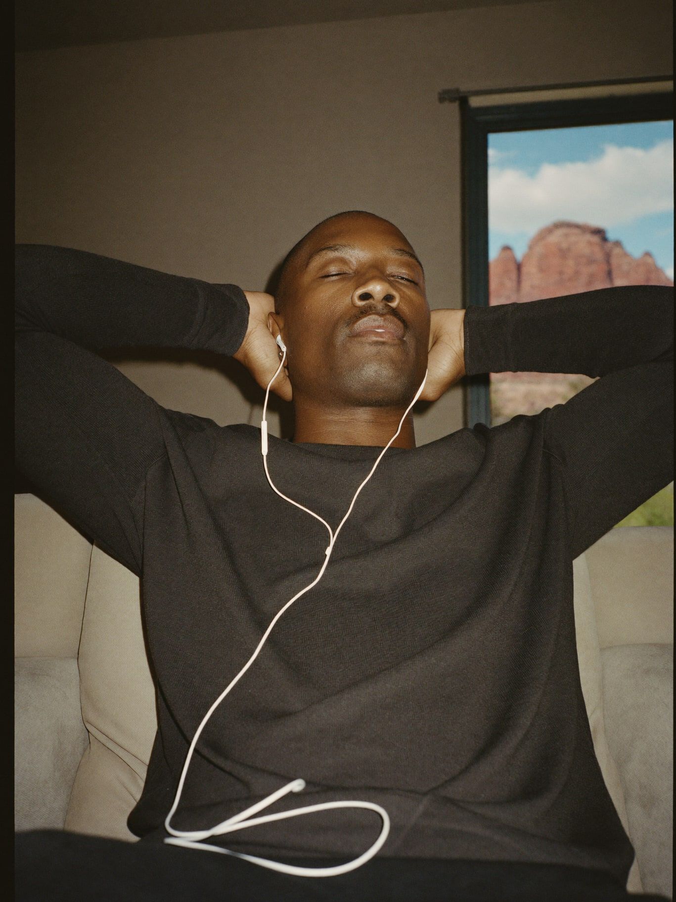 A Black man in his 40s wearing a long-sleeved charcoal sweater, sitting in a cozy armchair at home listening with earbuds, eyes closed and relaxed, a