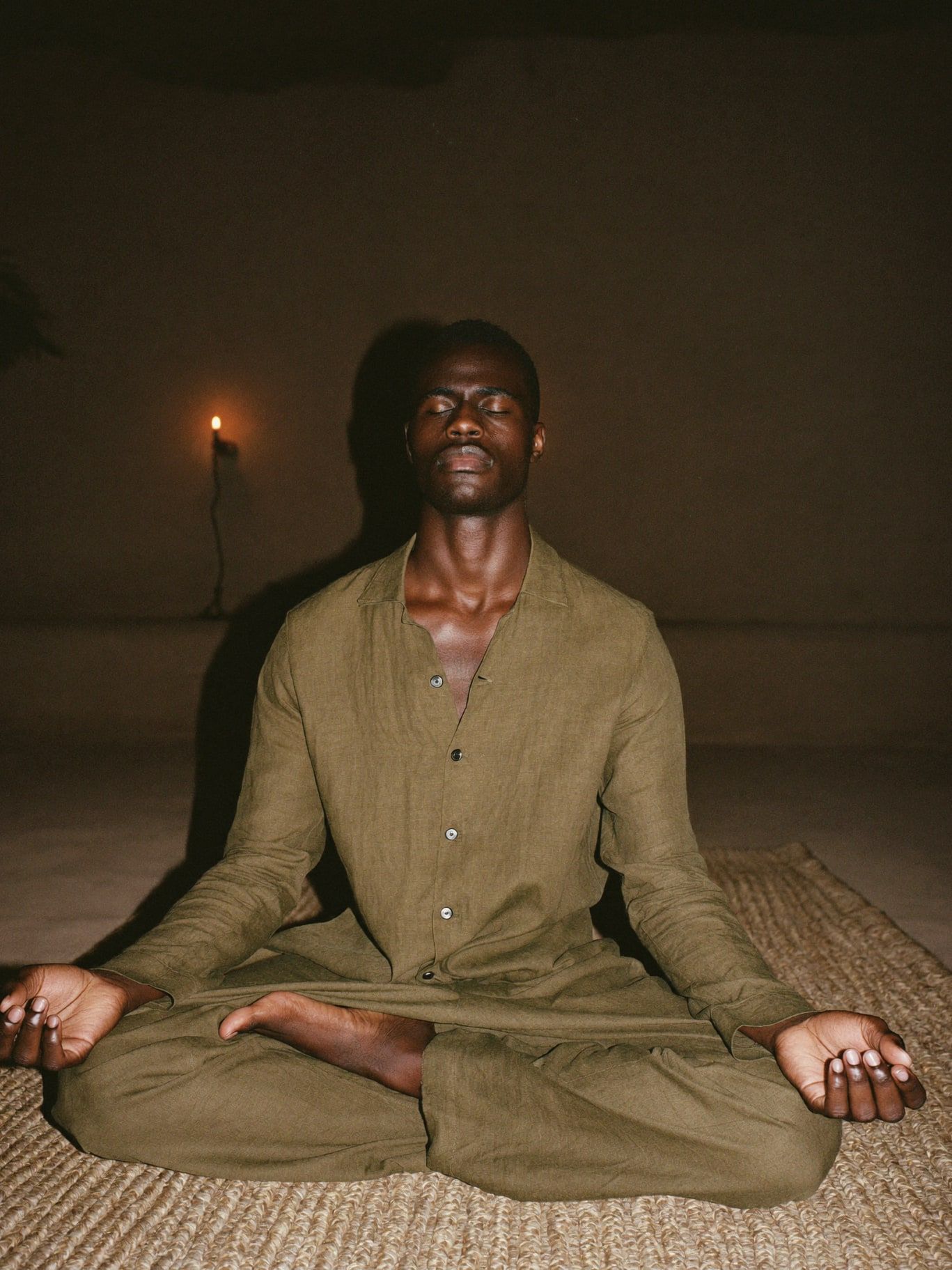 A Black man in his 30s wearing a buttoned long-sleeved olive linen shirt and loose trousers, meditating cross-legged on a woven rug in a dim adobe