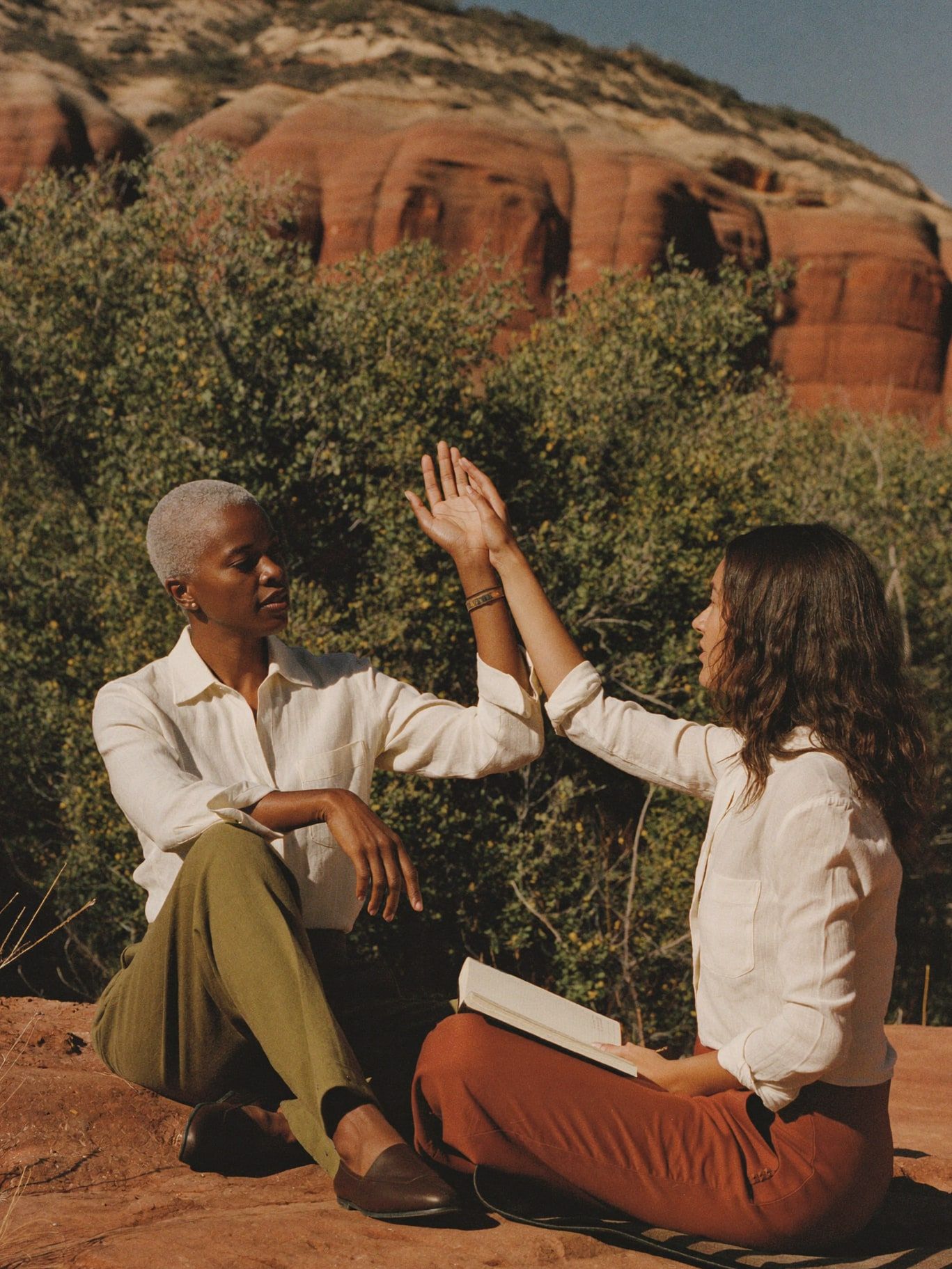 A trainer with short silver hair guiding a student's raised hand as they sit facing each other on red rock in Sedona