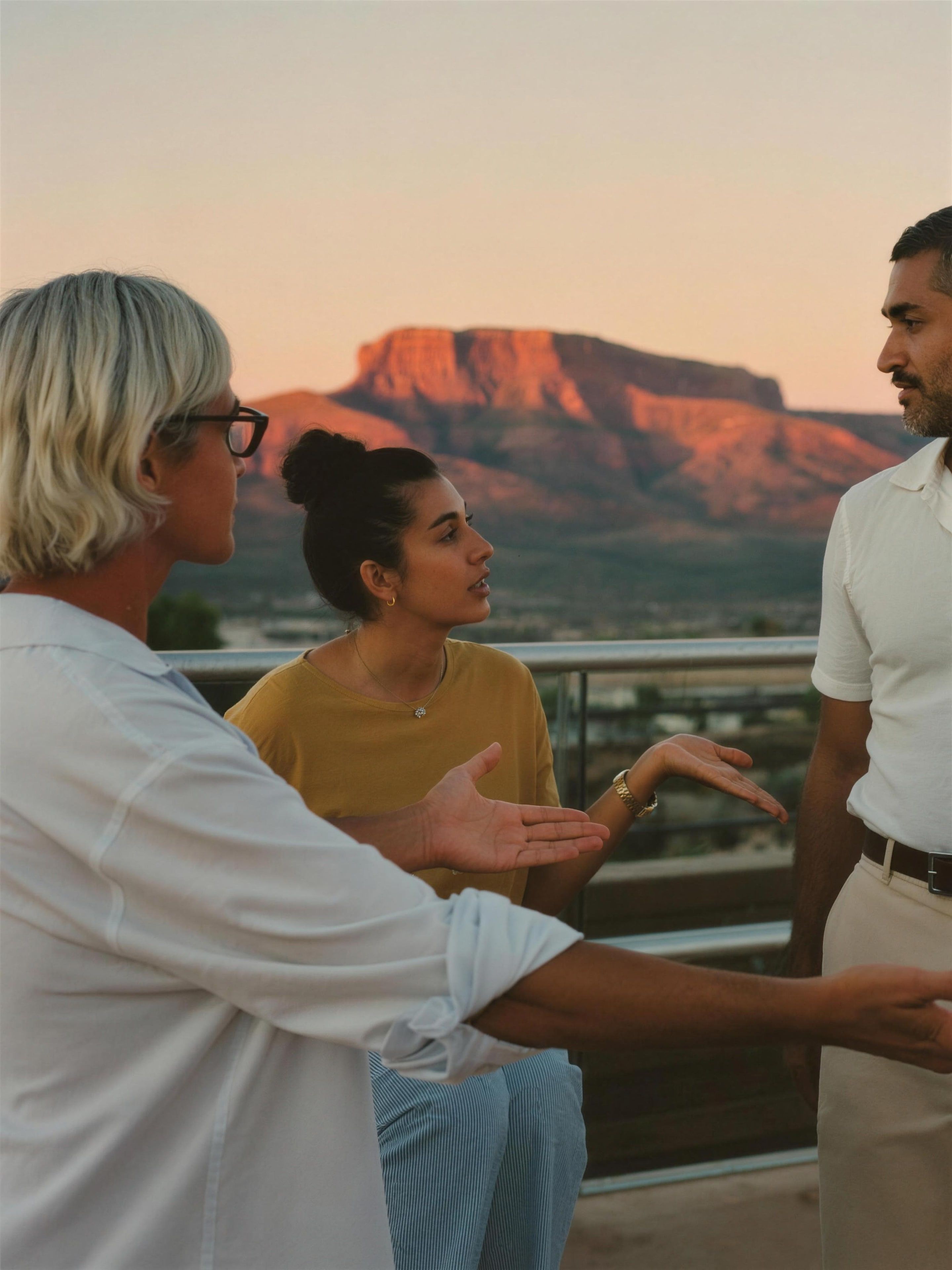 Three adults of color in quiet discussion on a terrace overlooking Sedona at golden hour, one speaking with open hands, the others listening