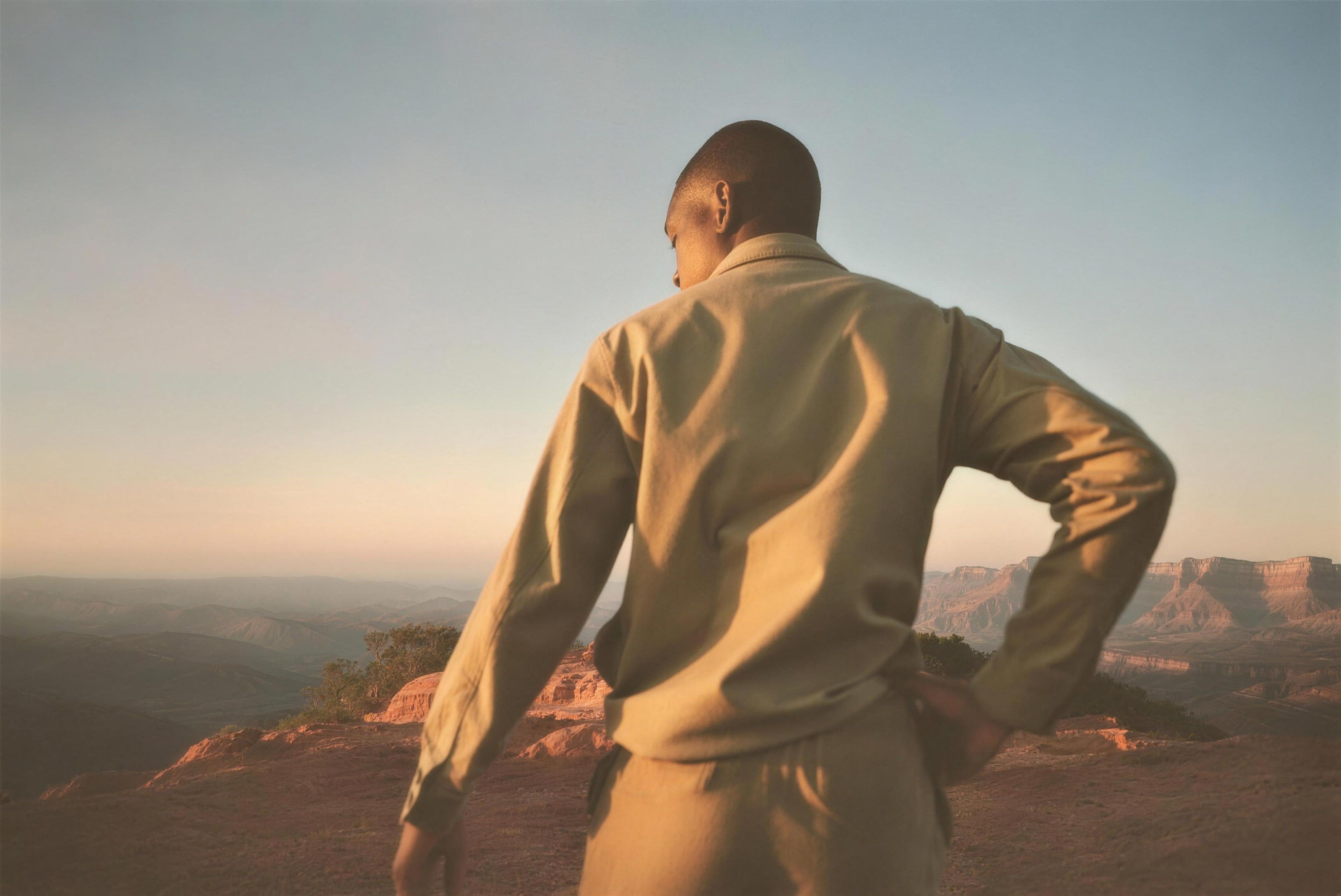 A Black man in his 40s wearing a long-sleeved olive jacket and trousers, seen from behind at a distance standing on a red rock overlook at sunrise
