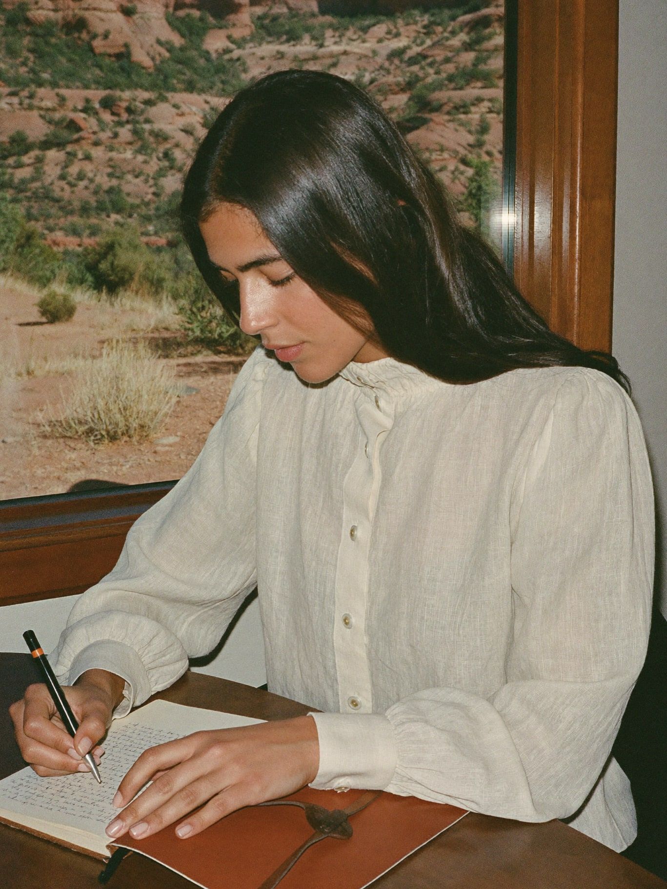 A woman of Middle Eastern descent in her 30s wearing a long-sleeved cream linen blouse, writing in a leather journal at a wooden desk by a window