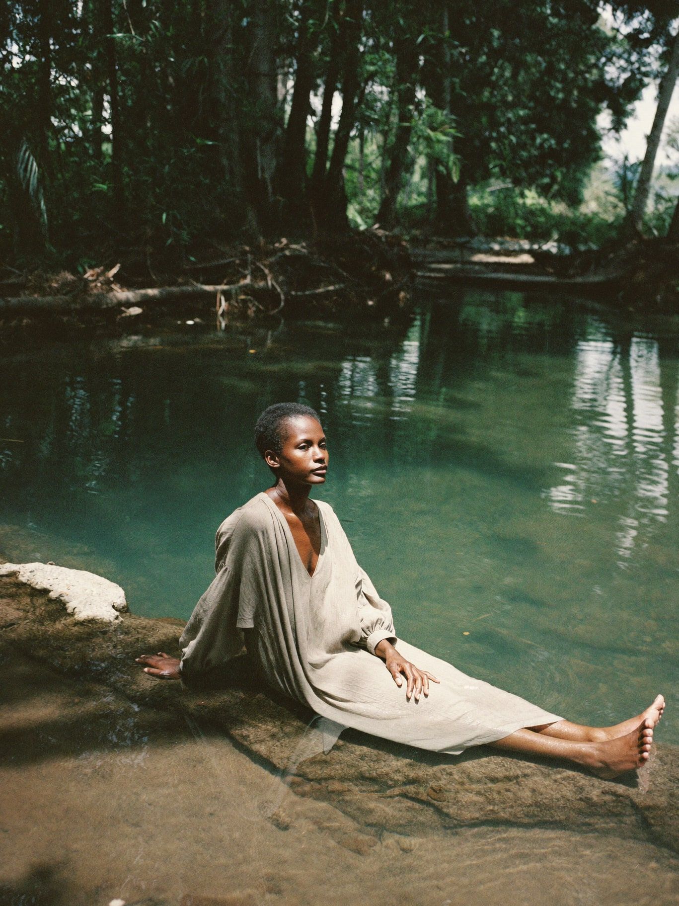 A Black woman in her 40s wearing a long loose linen dress, sitting at the edge of a clear jungle river pool in Honduras with her feet in the water