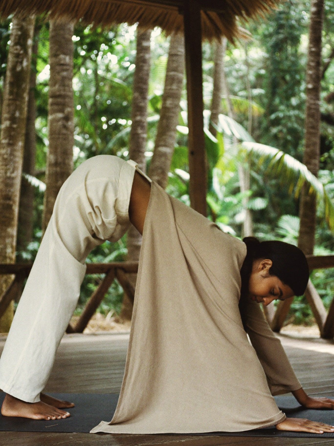 A South Asian woman wearing a long loose sand linen tunic that covers her waist and hips, and full-length linen trousers, practicing a gentle morning