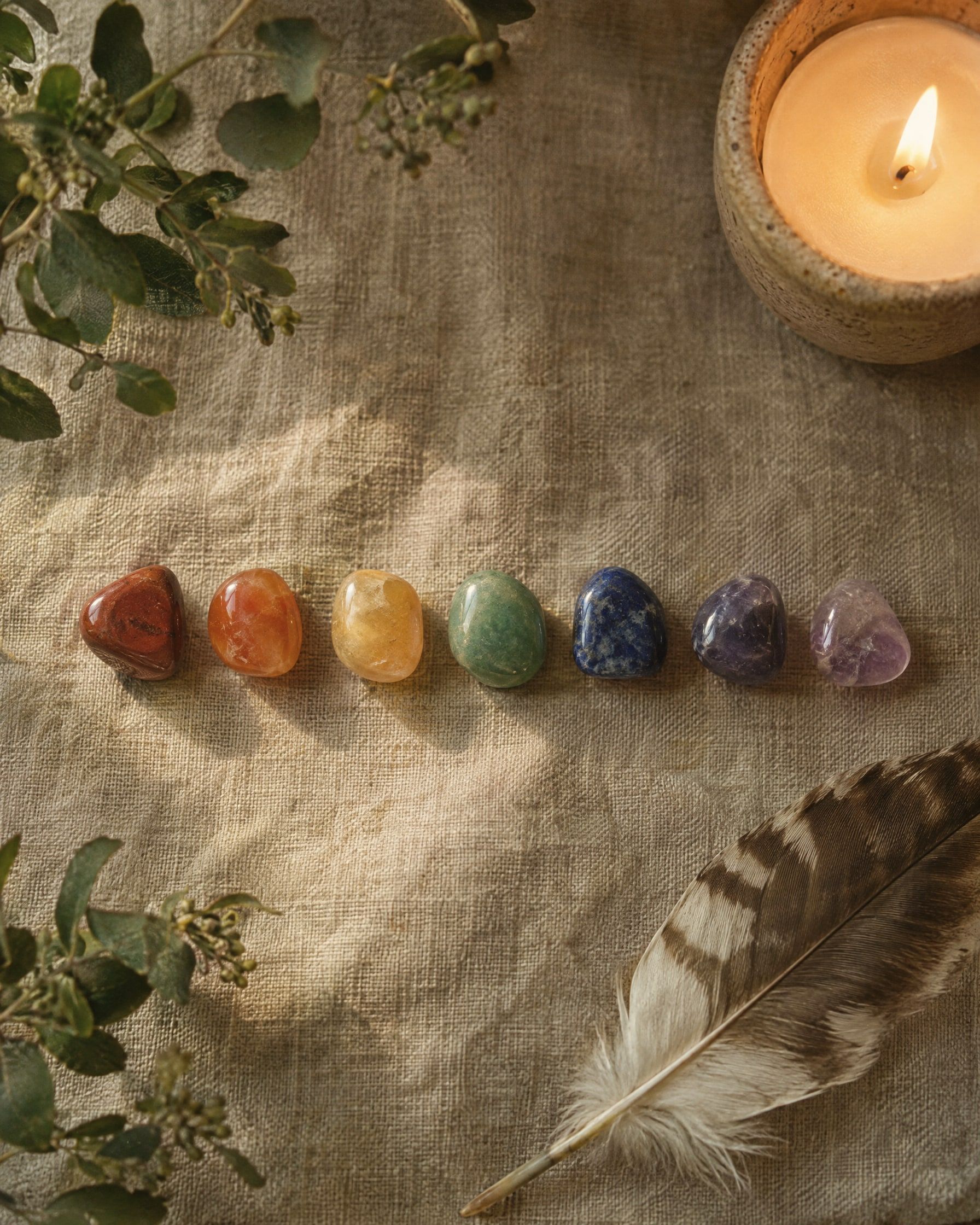 Seven small polished stones in earthy chakra tones of red, orange, yellow, green, blue, indigo and violet laid in a gentle line on raw linen beside a