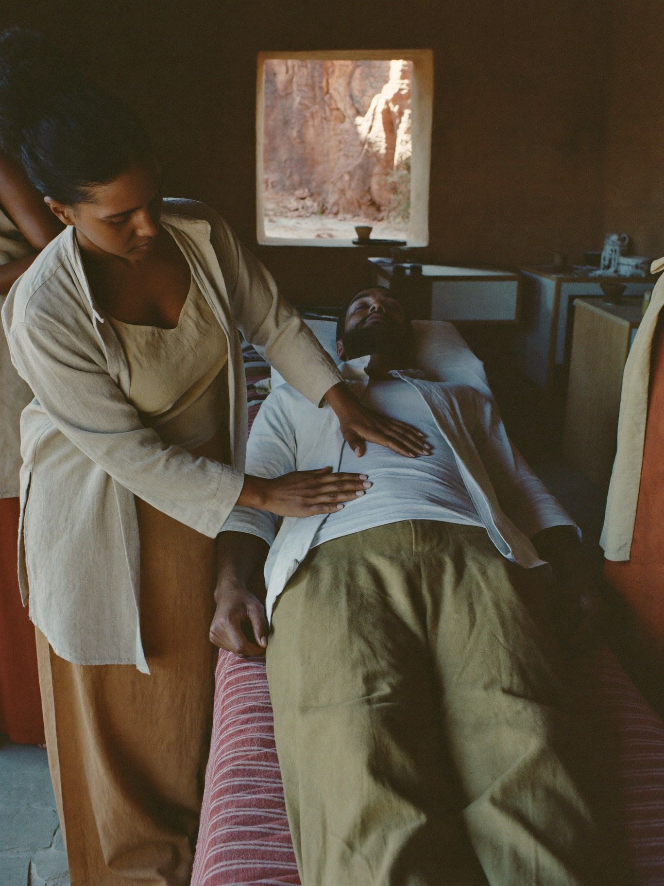 A Middle Eastern man in his 40s lying fully clothed on a linen-covered treatment table with eyes closed, a Black woman healer in a sand linen top