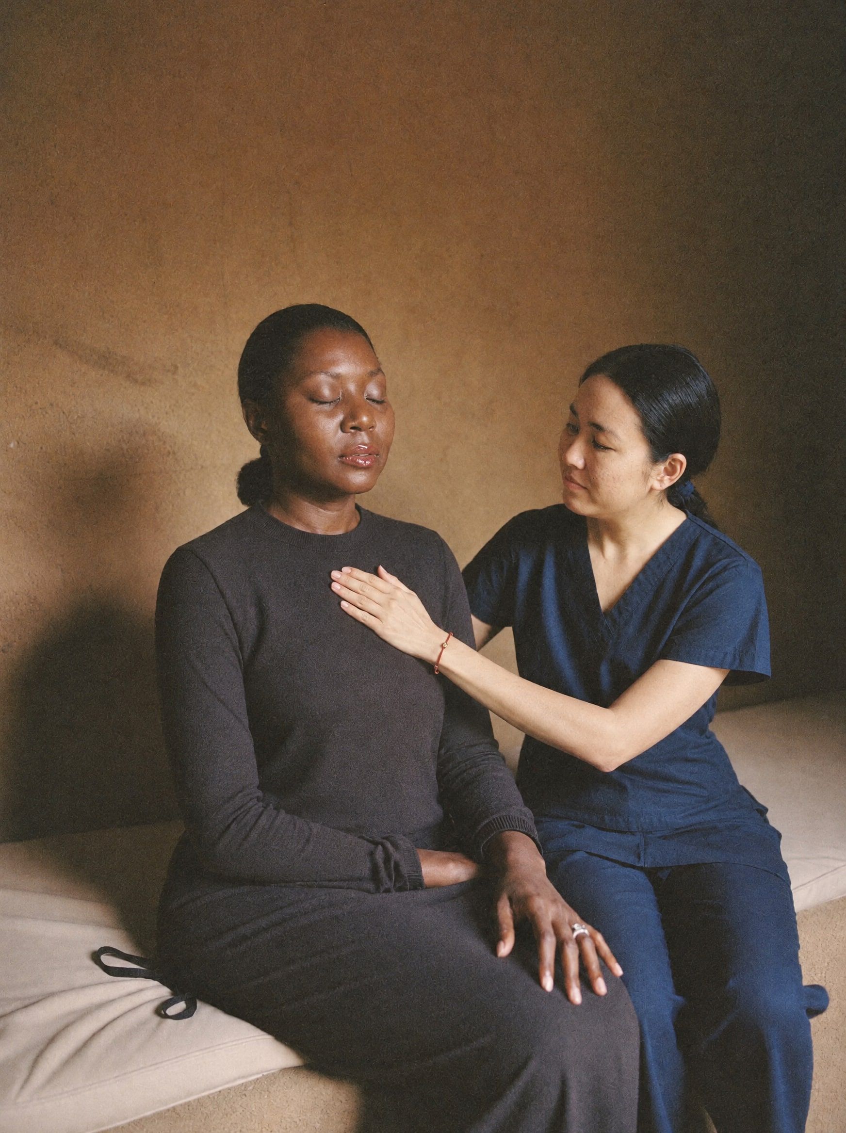 A Black woman in her 40s sitting on a cushioned bench with eyes closed, a Filipina practitioner beside her resting one hand on her upper chest at the heart center