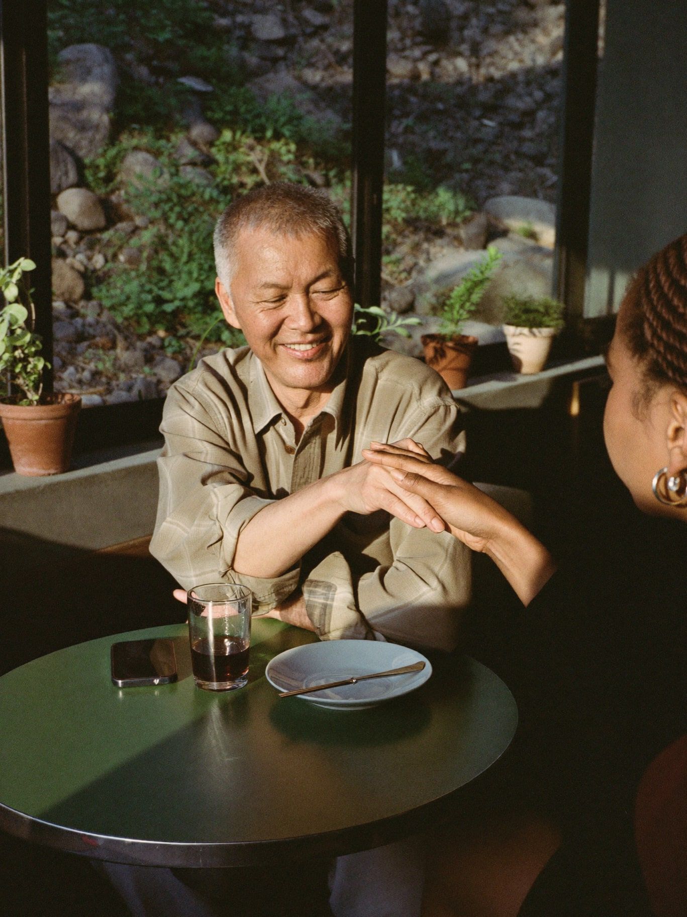 A Korean man in his 60s with short gray hair and a calm smile, wearing an olive linen shirt, reading the palm of a young Black woman with braids at a