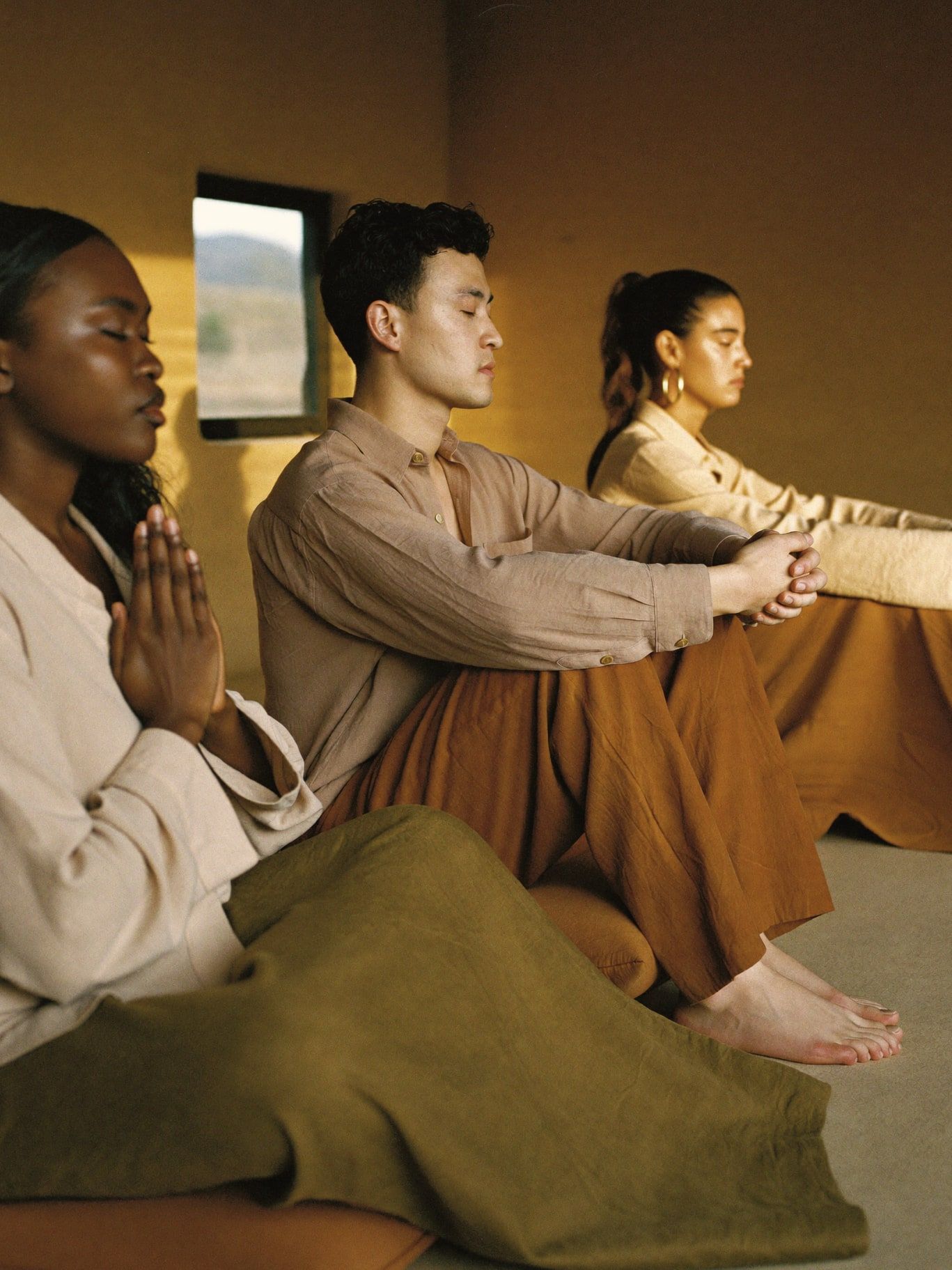 Three adults of color, a Black woman, a Korean man and a Latina woman, seated on floor cushions with eyes closed in a quiet adobe meditation room