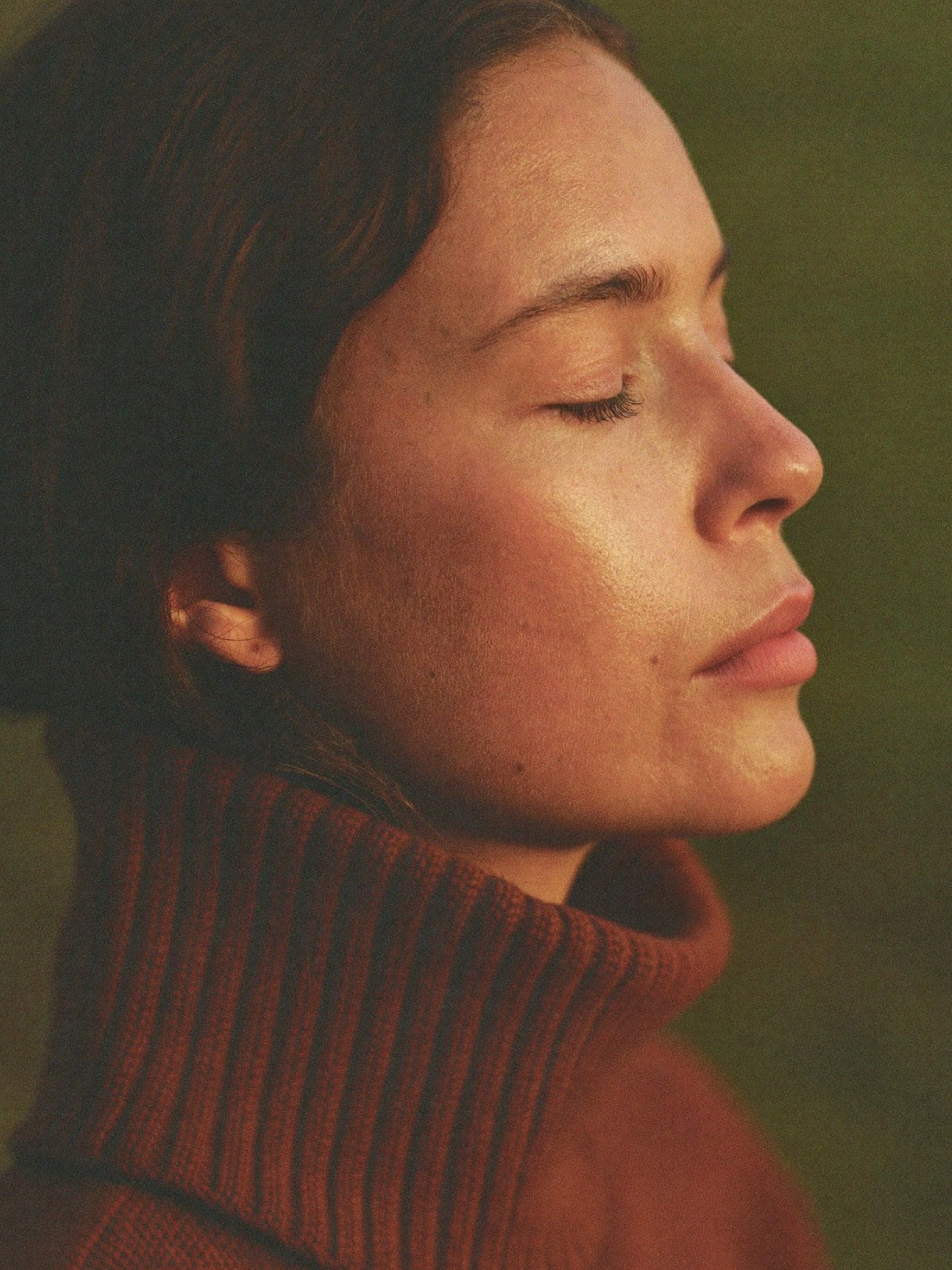 A Latina woman in her 30s wearing a high-necked rust knit sweater, eyes gently closed in meditation, warm dusk light falling across her face, deep
