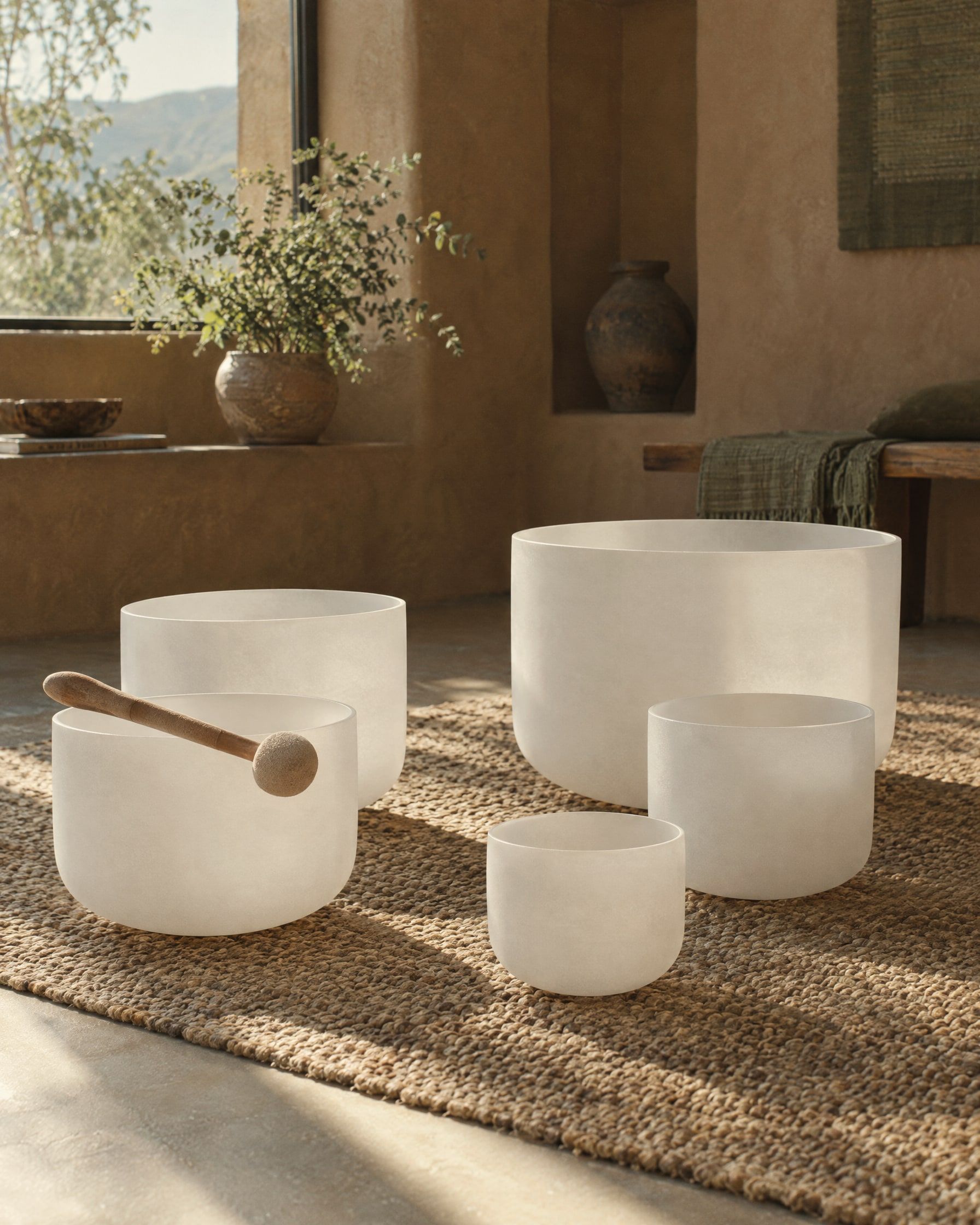 Frosted crystal singing bowls of different sizes arranged on a woven rug in a quiet adobe room, a suede mallet resting on one bowl, a sunbeam across