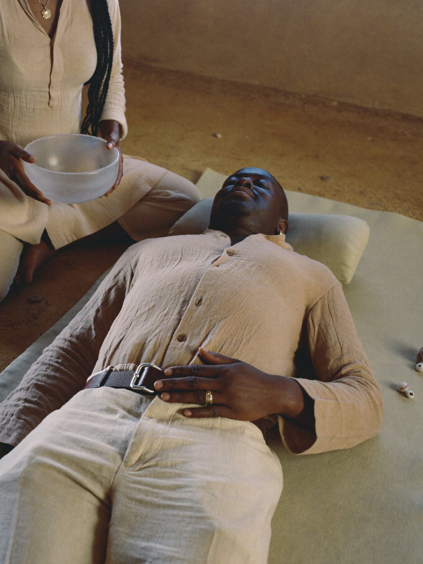 A Black man in his 40s wearing a long-sleeved sand linen shirt and linen trousers, fully clothed, lying on a cushioned mat with eyes closed while a