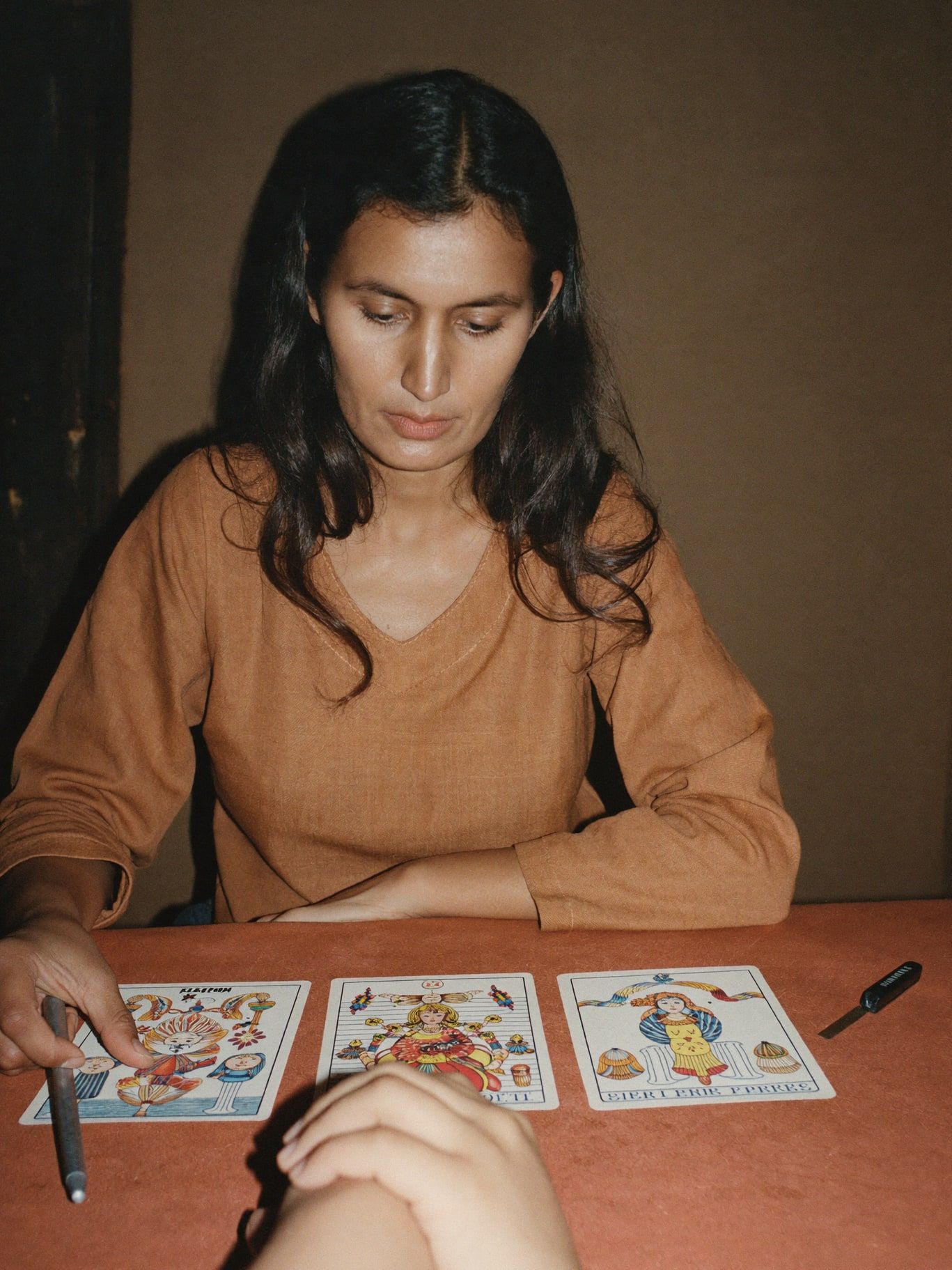 A South Asian woman in her 40s with long dark hair, wearing a rust linen top, laying three illustrated tarot cards on terracotta linen for a client