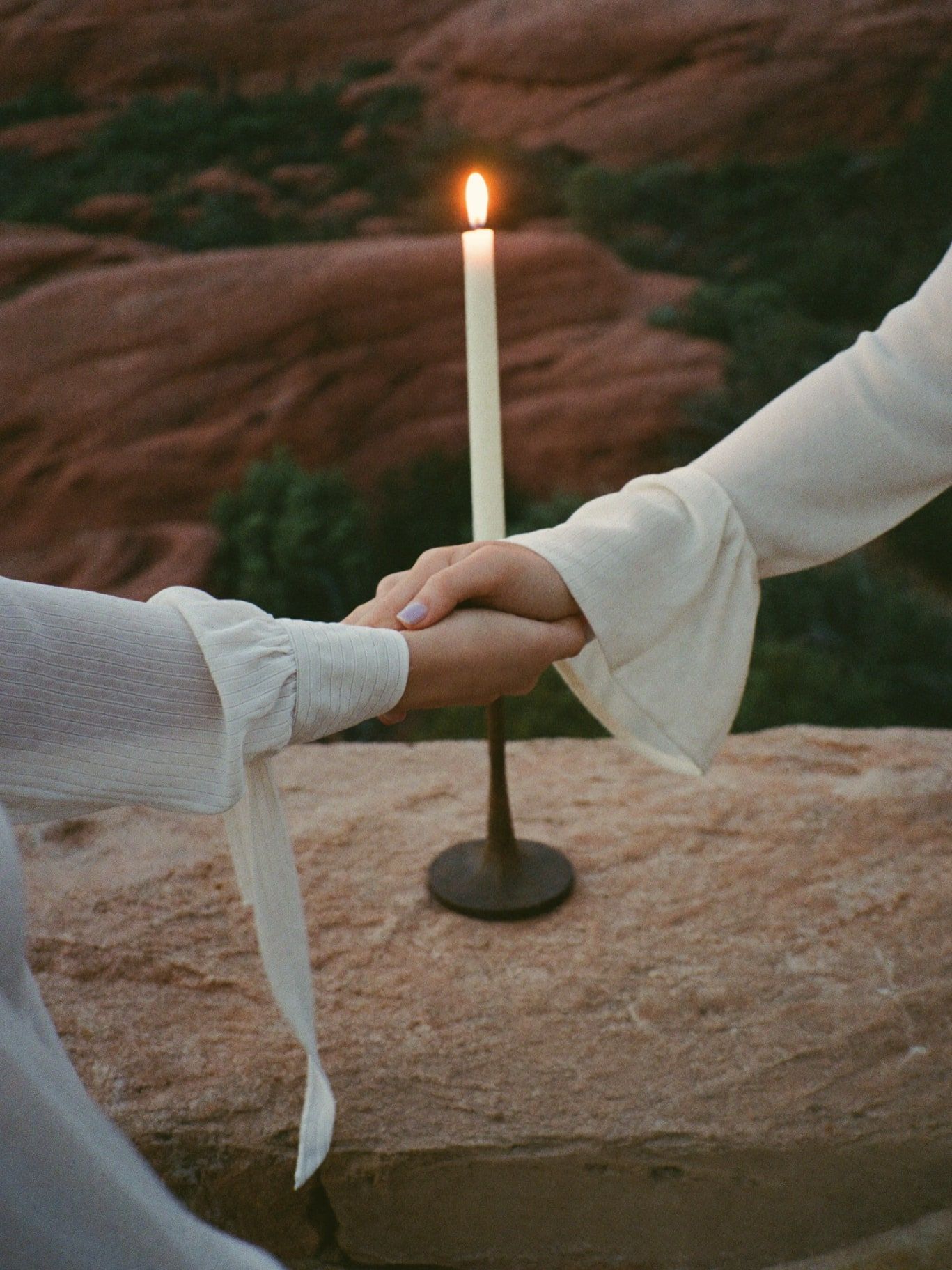 Three people's hands joined around a lit candle on a stone ledge at dusk in Sedona, long linen sleeves, warm candlelight, red rocks blurred behind