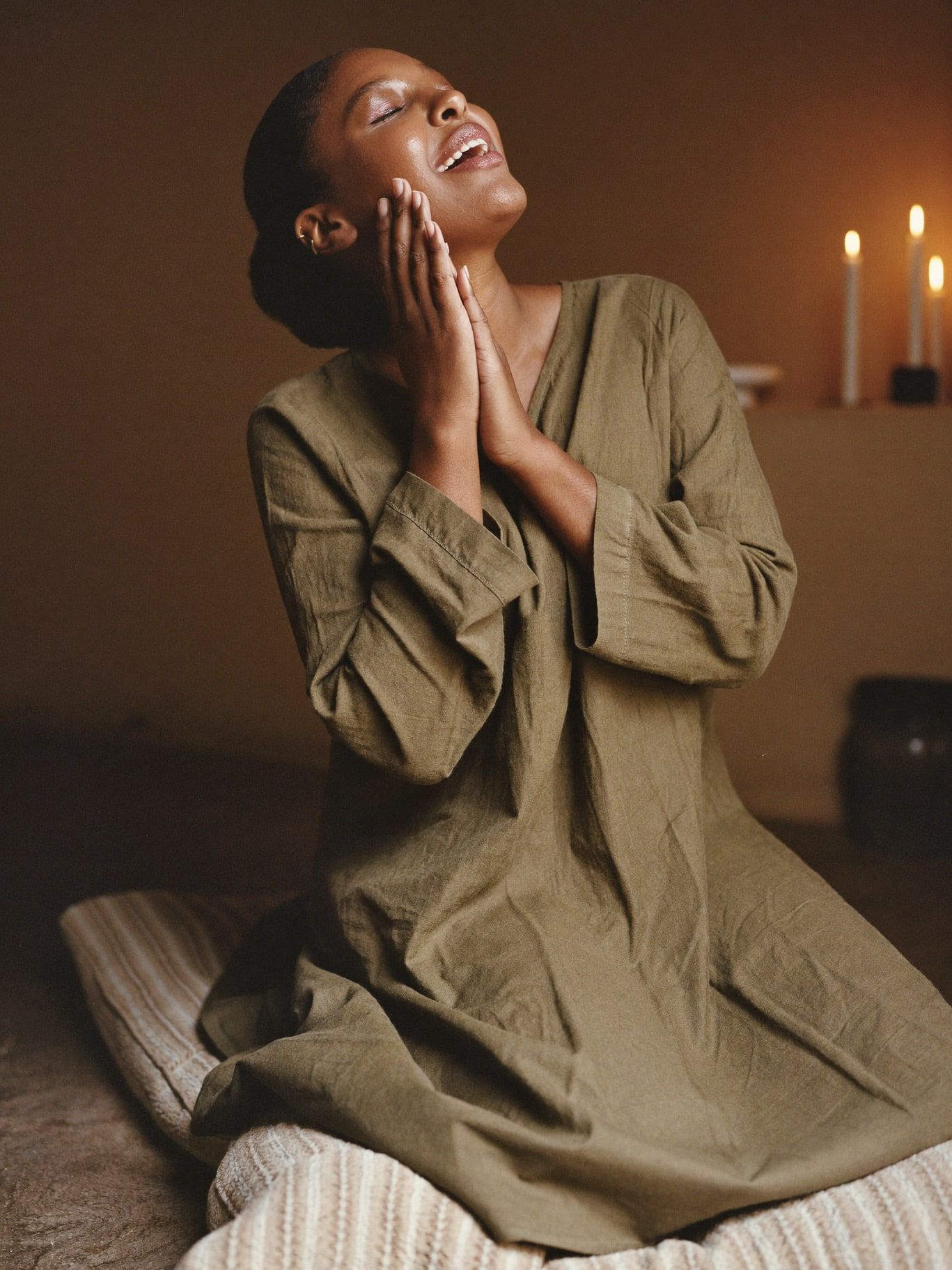 A Black woman in her 40s wearing a long-sleeved olive linen tunic, seated on a floor cushion with eyes closed, chanting softly with one hand at her